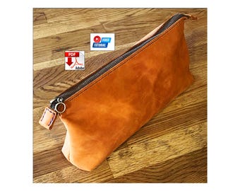 Può includere: Una pochette in pelle color cuoio con cerniera in metallo scuro, un tiralampo circolare in metallo e una piccola linguetta in pelle con cuciture blu. L'immagine include un'icona PDF Adobe e un'icona Video Tutorial blu e bianca.
