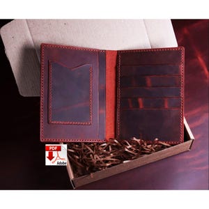 May include: Open, burgundy-colored leather wallet with multiple card slots and a pocket. The wallet is presented inside a cardboard box filled with brown packing material. The edges of the wallet are stitched with red thread.