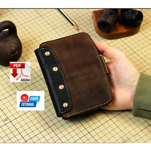 May include: A brown leather wallet with a black accent and copper rivets. The wallet is held in a hand, showcasing its design and craftsmanship. The background includes a weight, a pencil, and a cutting mat.