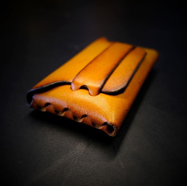 May include: A handcrafted, light brown leather wallet with a unique design. The wallet features a flap closure and decorative stitching along the edge. The leather has a rich, warm tone, and the wallet appears to be compact and durable.
