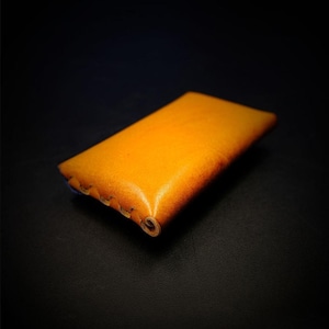 May include: A rectangular, handcrafted leather case in a warm, golden-yellow hue. The edges are stitched with a contrasting thread, and the leather has a slightly distressed appearance. The case is designed to hold small items.