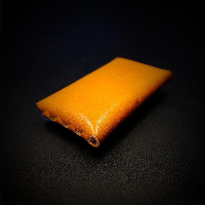 May include: A rectangular, handcrafted leather case in a warm, golden-yellow hue. The edges are stitched with a contrasting thread, and the leather has a slightly distressed appearance. The case is designed to hold small items.