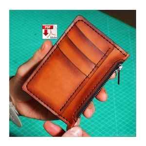 May include: A handcrafted, brown leather wallet with black stitching and a black zipper. The wallet has multiple card slots and is held in a person's hand. The Adobe PDF logo is in the upper left corner.