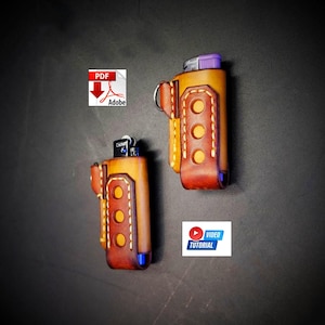 May include: Two leather lighter cases in a warm, golden-brown hue with contrasting dark brown accents. Each case features decorative stitching and a belt loop. One case has a Cricket lighter inside. The cases are displayed against a dark background.