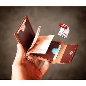 Könnte beinhalten: Ein braunes Lederportemonnaie mit einem Geldscheinclip, geöffnet, um Bargeld und eine Kreditkarte zu zeigen. Das Portemonnaie wird offen gehalten und zeigt sein Innendesign und die darin enthaltenen Gegenstände. Das Portemonnaie hat eine satte braune Farbe.