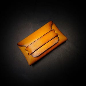 May include: A handcrafted, amber-colored leather cardholder with a secure flap closure. The wallet features a minimalist design with two horizontal straps and a dark brown edge. The leather has a natural grain and a warm, inviting tone.