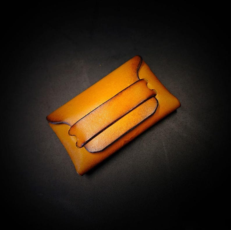 May include: A handcrafted, amber-colored leather cardholder with a secure flap closure. The wallet features a minimalist design with two horizontal straps and a dark brown edge. The leather has a natural grain and a warm, inviting tone.