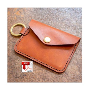 May include: A small, brown leather cardholder with a gold-colored metal ring attached. The cardholder has a flap secured by a gold snap. The edges are stitched with dark thread. The item is likely handmade.
