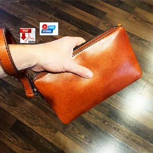 May include: A rectangular tan leather clutch bag featuring a matching wrist strap, a dark zip closure along the top edge, and visible black stitching along the seams. The surface of the leather has a smooth, polished finish with subtle colour variations.