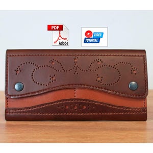 May include: A brown leather wallet with a decorative cut-out design on the flap. The wallet has a two-tone colour scheme, with a lighter brown section at the bottom. Two silver-coloured snaps secure the wallet. The wallet is on a wooden surface.