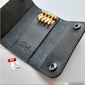 Puede incluir: Un llavero de cuero negro con aros dorados. El llavero está abierto, revelando el interior y los aros. La palabra "Handy" está grabada en el cuero. Se ven dos cierres de botón plateados.