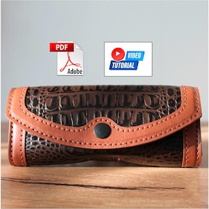 Puede incluir: Cartera de cuero marrón con estampado de cocodrilo, ribete de cuero marrón claro y cierre de botón negro. La imagen también incluye iconos para un documento PDF y un tutorial en vídeo.