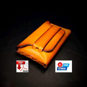 Può includere: Un portafoglio in pelle arancione fatto a mano con chiusura a patta. Il portafoglio ha un colore caldo e una consistenza liscia. Il design include una patta decorativa e un bordo marrone scuro a contrasto. L'immagine include anche un'icona di tutorial PDF e video.