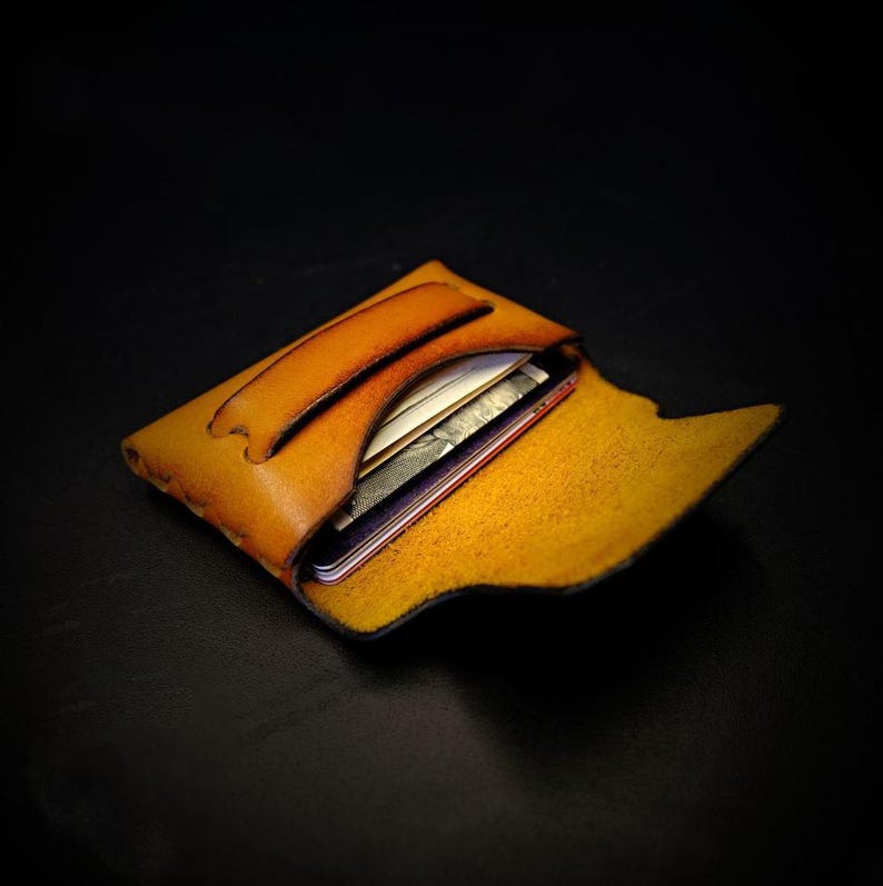 May include: A handcrafted, open leather wallet in a warm, golden-brown hue. The wallet features a curved handle and is filled with cards and a smartphone. The interior is a lighter shade of yellow. The wallet is designed to hold essentials.