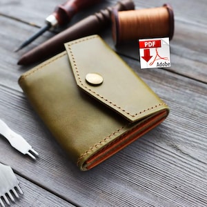 Op de afbeelding: Olijfgroene leren portemonnee met een klepsluiting en een goudkleurige drukknop. De portemonnee heeft een oranje binnenvoering. Leren gereedschappen, waaronder een naald, draad en een stikgereedschap, zijn zichtbaar.