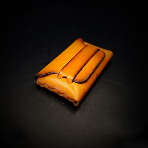 May include: A handcrafted, rectangular leather wallet in shades of tan and brown. The wallet features a flap closure with a tab and a decorative, stitched edge. The leather has a natural, textured appearance, suggesting quality craftsmanship.