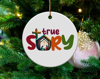 Puede incluir: Un adorno navideño redondo y blanco con las palabras "true story" en letras coloridas. El adorno presenta una cruz, un pesebre y está colgado de un árbol de Navidad.