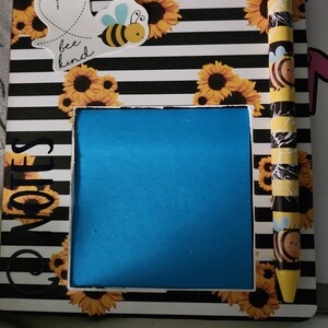 May include: A black and white striped notepad with a blue sticky note, a bee and sunflower design, and the text "bee kind". The notepad has a pen with a bee and sunflower design.