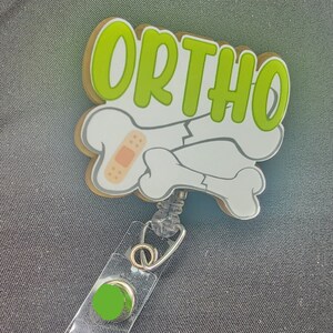 Könnte beinhalten: Ein Ausweis-Rollhalter mit dem Wort "ORTHO" in Grün, umrandet von Weiß und Beige, über einer Grafik von zwei weißen Knochen mit einem Pflaster. Die Rolle hat einen transparenten Kunststoffriemen und einen grünen Knopf.