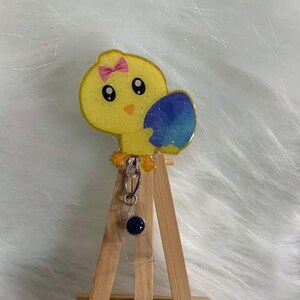 May include: A yellow chick with a pink bow holding a blue and green speckled egg. The chick is a resin charm attached to a retractable badge holder with a dark blue button.