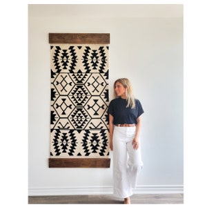 May include: A black and white geometric tapestry hanging on a wall with a wooden frame. A person is standing next to the tapestry wearing a black shirt and white pants.