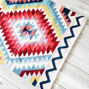 May include: A colourful geometric rug with a white background and a diamond pattern in shades of red, blue, yellow, and pink. The rug has a zig-zag border in navy blue.