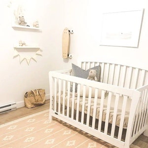 May include: A white cot with a white mattress and a white and grey striped pillow. A stuffed toy is sitting on the pillow. The cot is in a nursery with a white rug and a white wall. There is a skateboard hanging on the wall.