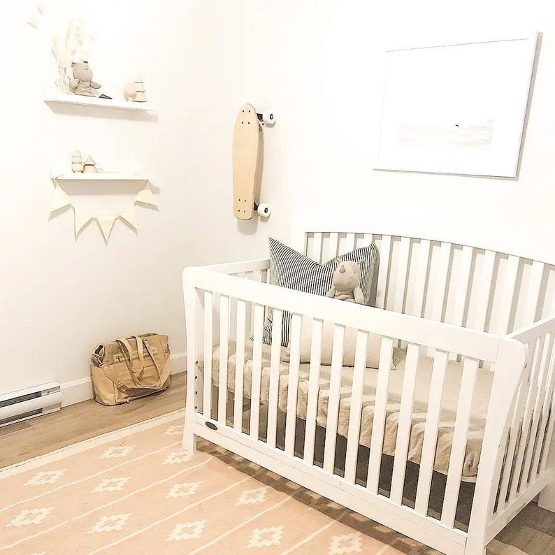 May include: A white cot with a white mattress and a white and grey striped pillow. A stuffed toy is sitting on the pillow. The cot is in a nursery with a white rug and a white wall. There is a skateboard hanging on the wall.