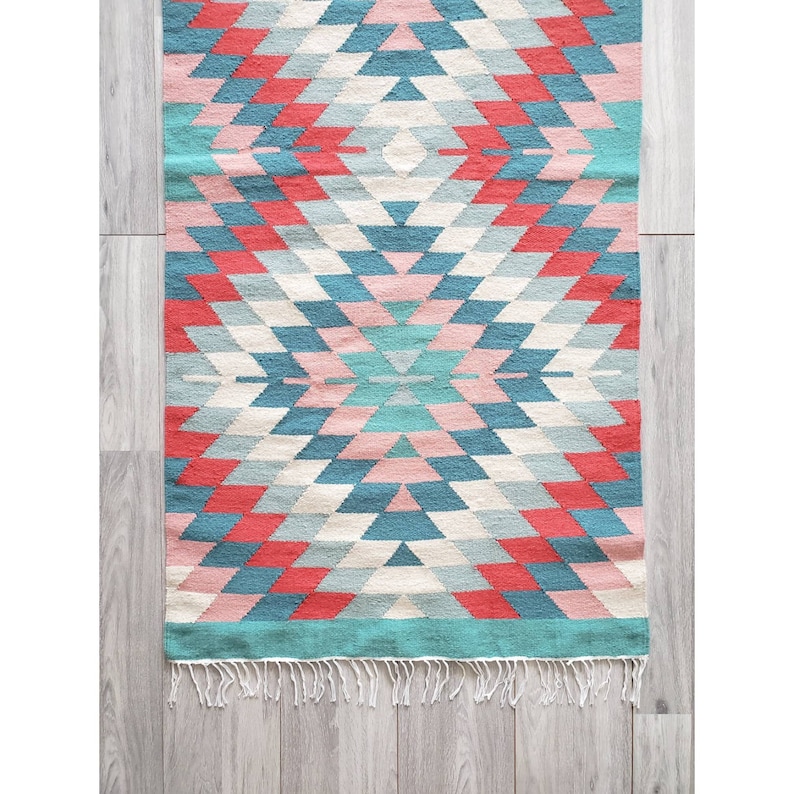 May include: A colorful geometric rug with a diamond pattern in shades of pink, blue, and red. The rug has a fringe border.