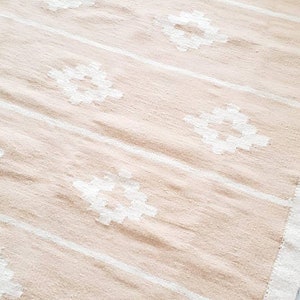 May include: A beige and white woven rug with a geometric pattern. The rug has a fringed border.