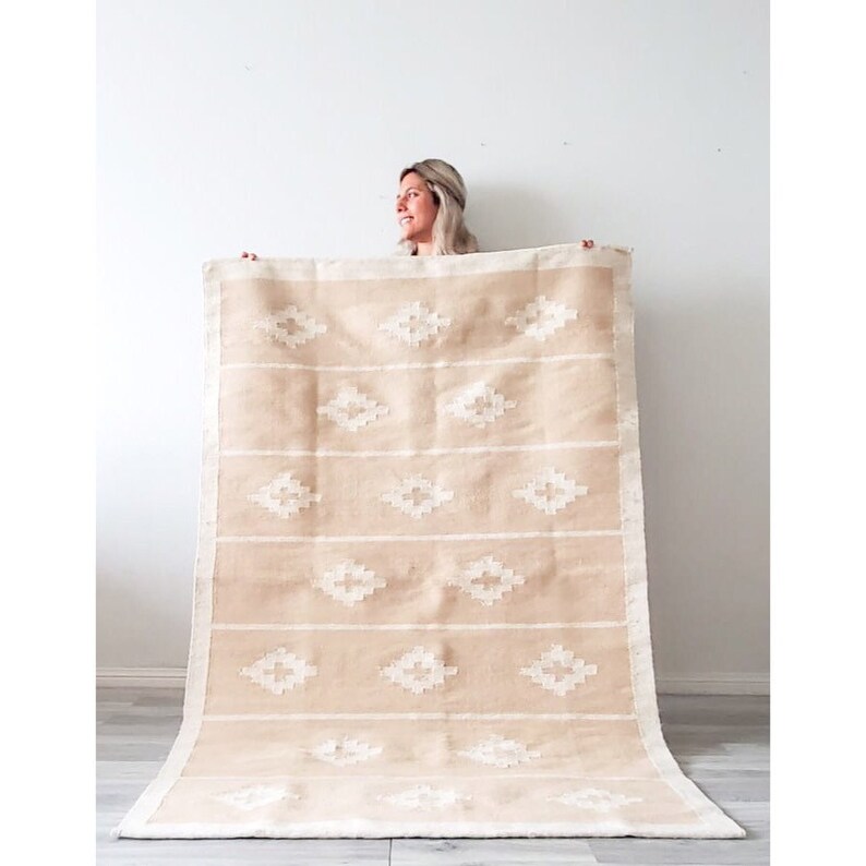 May include: A beige and white woven rug with a geometric diamond pattern. The rug is being held up by a person.
