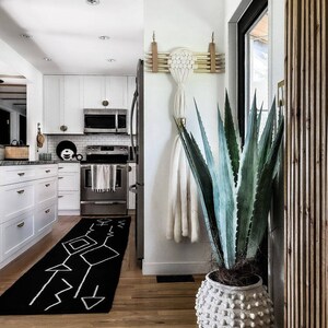 May include: A white planter with a textured surface holds a large green agave plant. The plant is in the corner of a room with a white wall and a wooden door. A black and white geometric rug is on the floor.