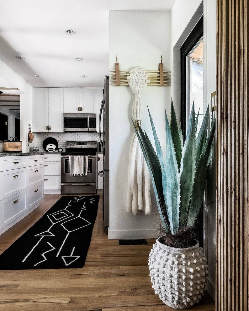 May include: A white planter with a textured surface holds a large green agave plant. The plant is in the corner of a room with a white wall and a wooden door. A black and white geometric rug is on the floor.