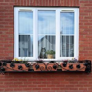 Könnte beinhalten: Ein Blumenkasten mit bunten Blumen und Grünpflanzen befindet sich unter einem weiß gerahmten Fenster an einer roten Backsteinmauer. Der Blumenkasten ist aus schwarzem Metall mit dekorativen Blättern und terrakottafarbenen Töpfen.