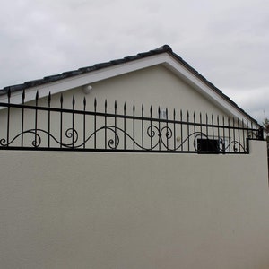 Puede incluir: Una pared blanca con una valla de metal negro con volutas decorativas y puntas. Una puerta blanca con un marco de metal negro y un diseño floral negro se encuentra en el lado derecho de la imagen.