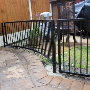 Puede incluir: Valla de metal negro con una secci&oacute;n curva y una puerta. La valla est&aacute; en un patio trasero con una terraza y c&eacute;sped.