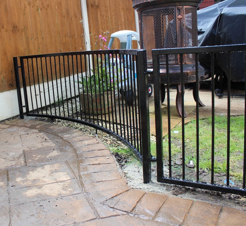 Puede incluir: Valla de metal negro con una secci&oacute;n curva y una puerta. La valla est&aacute; en un patio trasero con una terraza y c&eacute;sped.