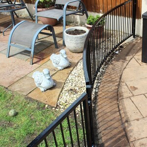 Puede incluir: Valla curva de metal negro con barras verticales, instalada en un patio trasero. La valla est&aacute; parcialmente oculta por una planta en maceta y dos figuras decorativas de patos.