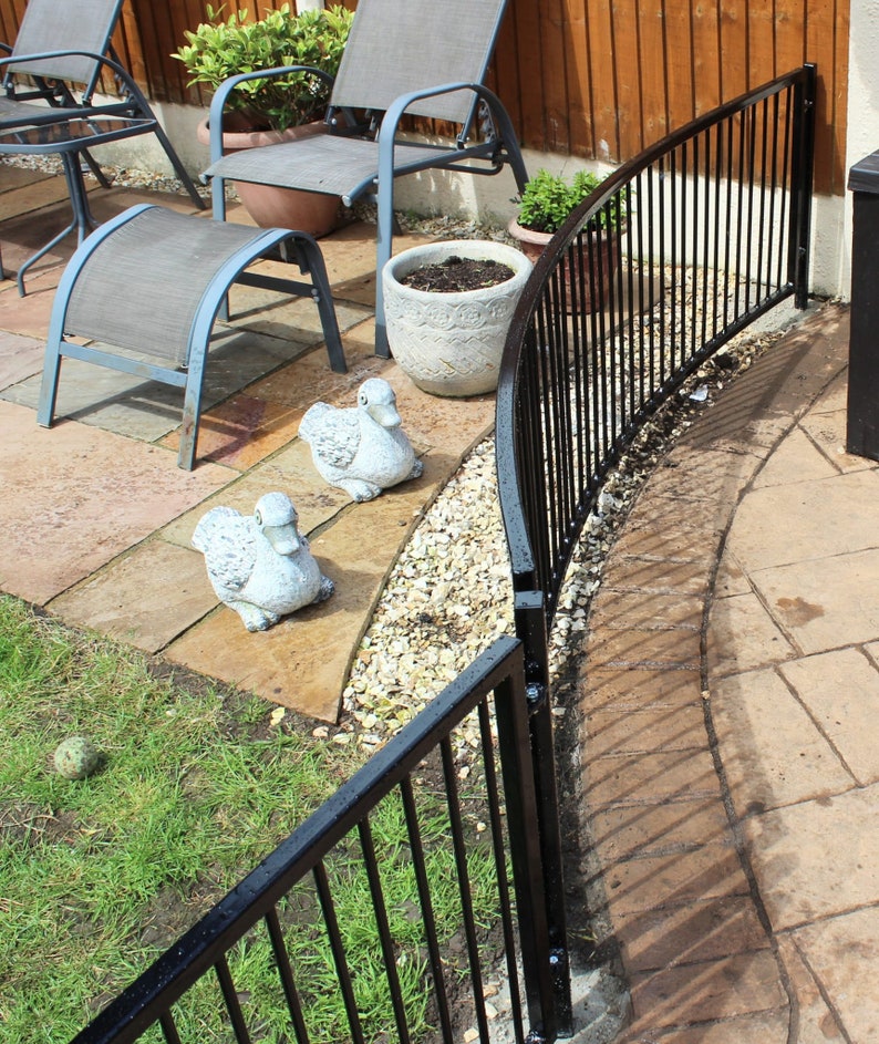 Puede incluir: Valla curva de metal negro con barras verticales, instalada en un patio trasero. La valla est&aacute; parcialmente oculta por una planta en maceta y dos figuras decorativas de patos.