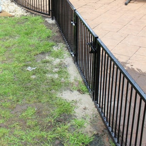 Puede incluir: Valla de metal negro con una puerta, instalada en un jard&iacute;n. La valla es curva y tiene barras verticales. La puerta est&aacute; abierta y tiene un pestillo.