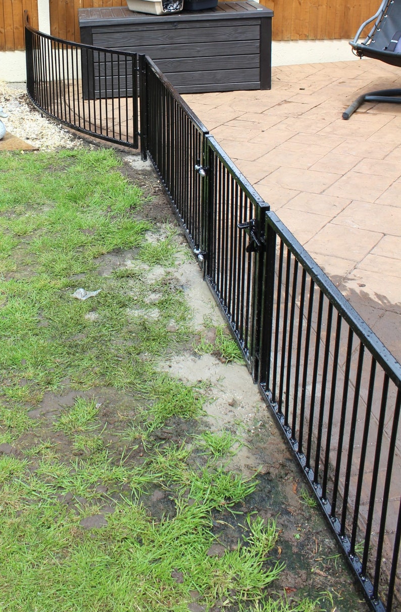 Puede incluir: Valla de metal negro con una puerta, instalada en un jard&iacute;n. La valla es curva y tiene barras verticales. La puerta est&aacute; abierta y tiene un pestillo.