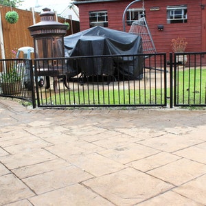 Puede incluir: Una valla de metal negro con una puerta en el medio, que separa una zona de patio de un jard&iacute;n trasero con c&eacute;sped. El patio est&aacute; hecho de adoquines de hormig&oacute;n y hay una barbacoa cubierta en el fondo.