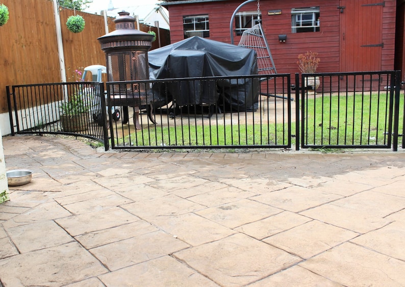 Puede incluir: Una valla de metal negro con una puerta en el medio, que separa una zona de patio de un jard&iacute;n trasero con c&eacute;sped. El patio est&aacute; hecho de adoquines de hormig&oacute;n y hay una barbacoa cubierta en el fondo.