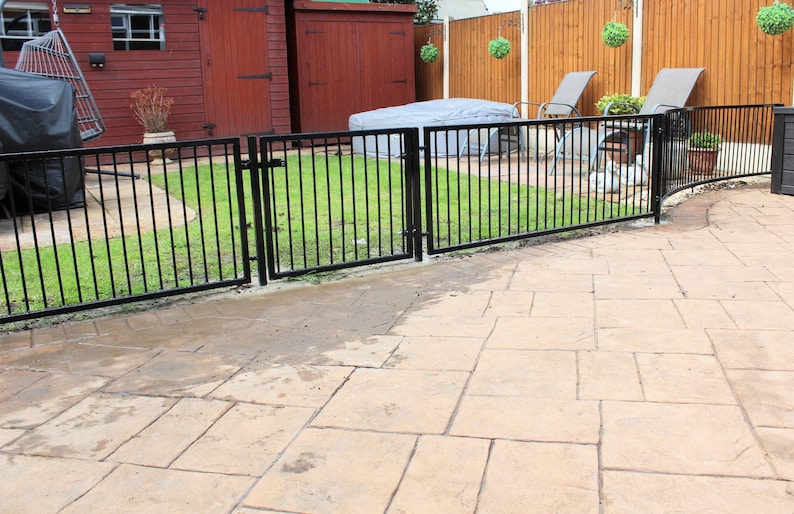 Puede incluir: Una valla de metal negro con dos puertas rodea un &aacute;rea de c&eacute;sped. La valla est&aacute; curvada en el extremo y tiene un patio de concreto en frente.