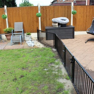 Puede incluir: Una valla de metal negro con una puerta en un patio trasero. La valla es curva y tiene una valla de madera detr&aacute;s. Hay una caja de almacenamiento marr&oacute;n y un c&eacute;sped verde en el fondo.