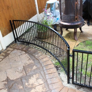 Puede incluir: Paneles de valla curvos de metal negro con una puerta. La valla est&aacute; instalada en un patio trasero con un patio de piedra y c&eacute;sped verde.