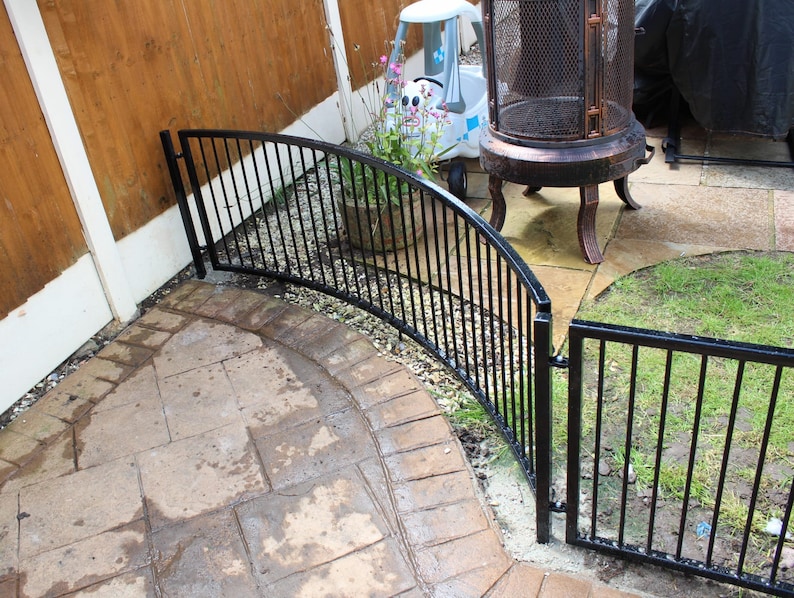 Puede incluir: Paneles de valla curvos de metal negro con una puerta. La valla est&aacute; instalada en un patio trasero con un patio de piedra y c&eacute;sped verde.