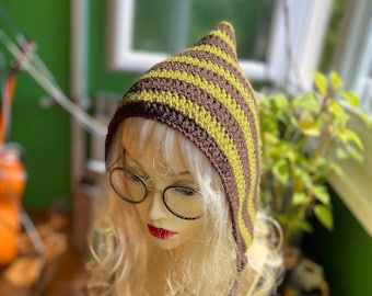 May include: A brown and green striped crocheted hat with a pointed hood and long ties. The hat is on a mannequin with long blonde hair.
