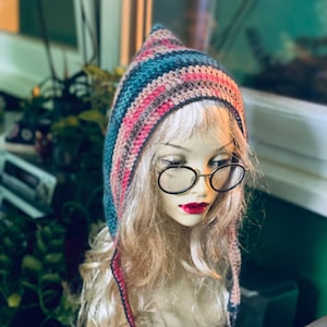 Peut inclure: Un chapeau crocheté multicolore avec un sommet pointu et des attaches d'oreille. Le chapeau présente des rayures roses, bleues et beiges. Il est présenté sur une tête de mannequin avec des cheveux blonds, des lunettes et du rouge à lèvres rouge.