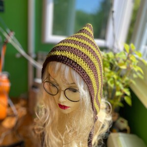 Può includere: Un cappello a maglia a strisce marroni e verdi con un cappuccio a punta e lunghi lacci. Il cappello è su un manichino con lunghi capelli biondi.